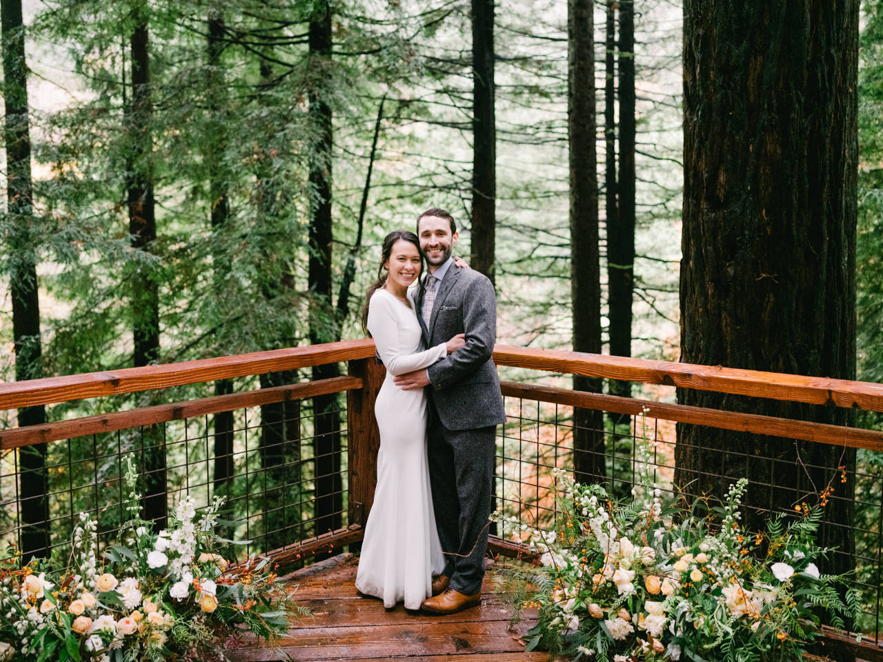  Bride and groom pose on redwood deck in Hoyt arboretum after wedding ceremony with large floral centerpieces on each side 