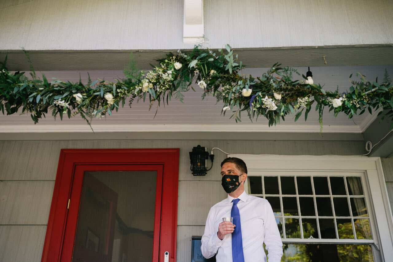  Groom in floral mask waits on front porch by red door 