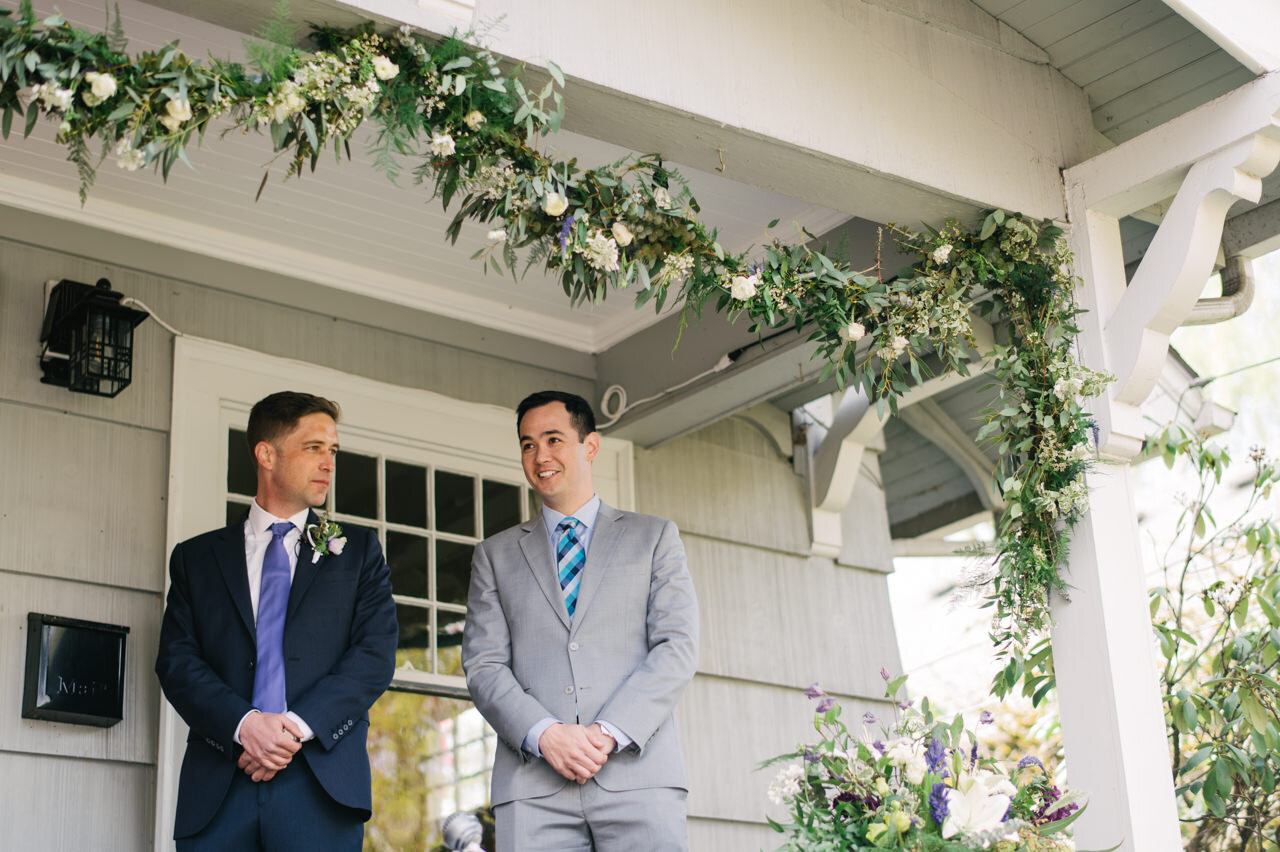  Groom in blue and purple suit and best man in grey and light blue tie wait under floral string on front porch elopement to begin 