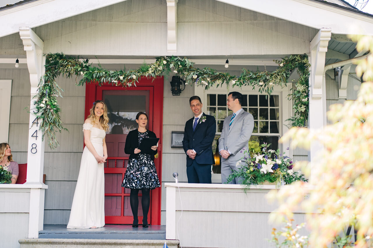  Bride and groom stand together on front porch in front of red door looking out on street during wedding elopement 