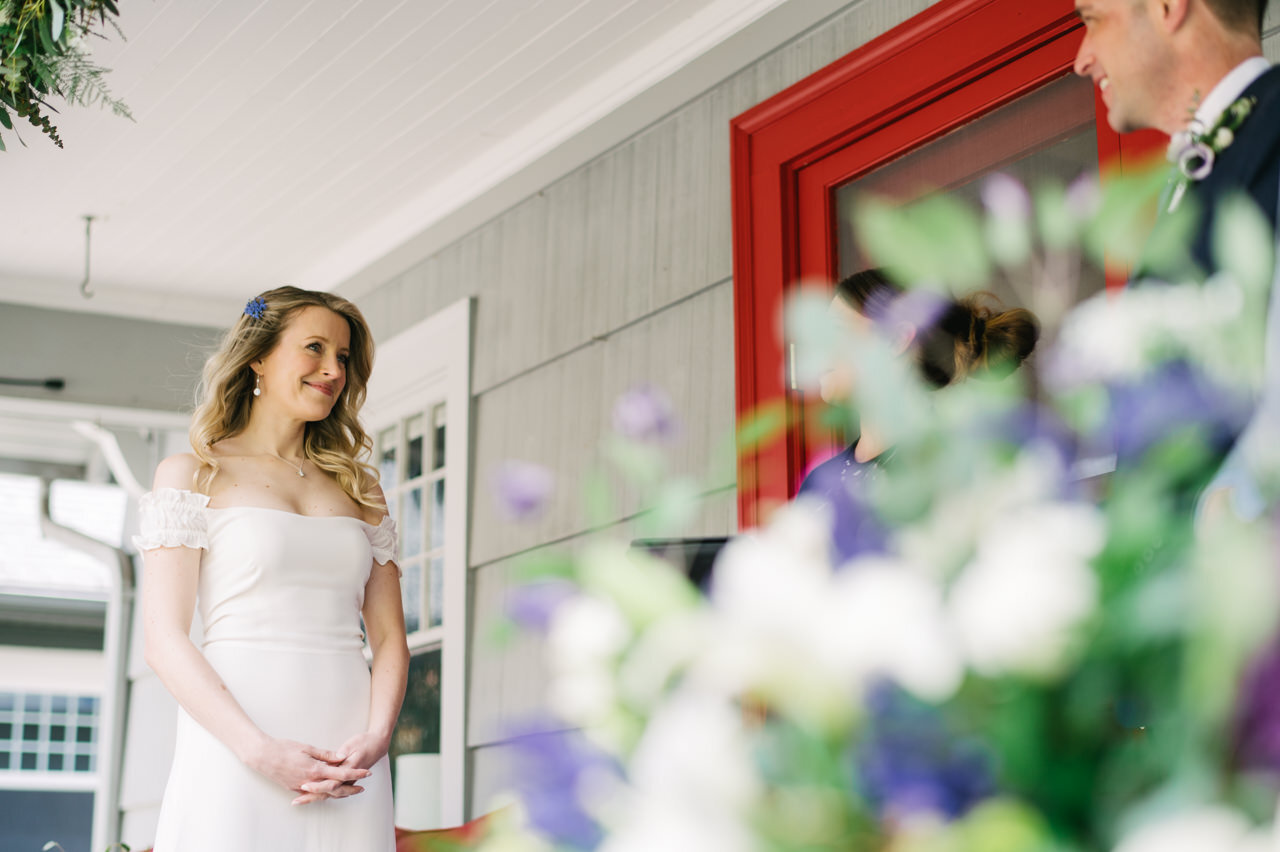 Bride smiles at groom on front porch with purple and white flowers in the foreground 