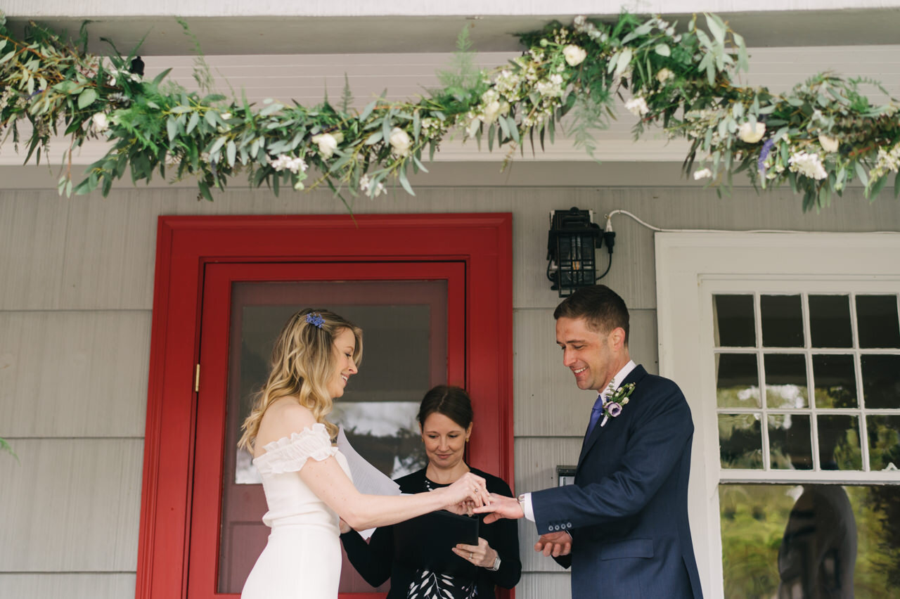  Bride places ring on groom's finger in front of home red door 