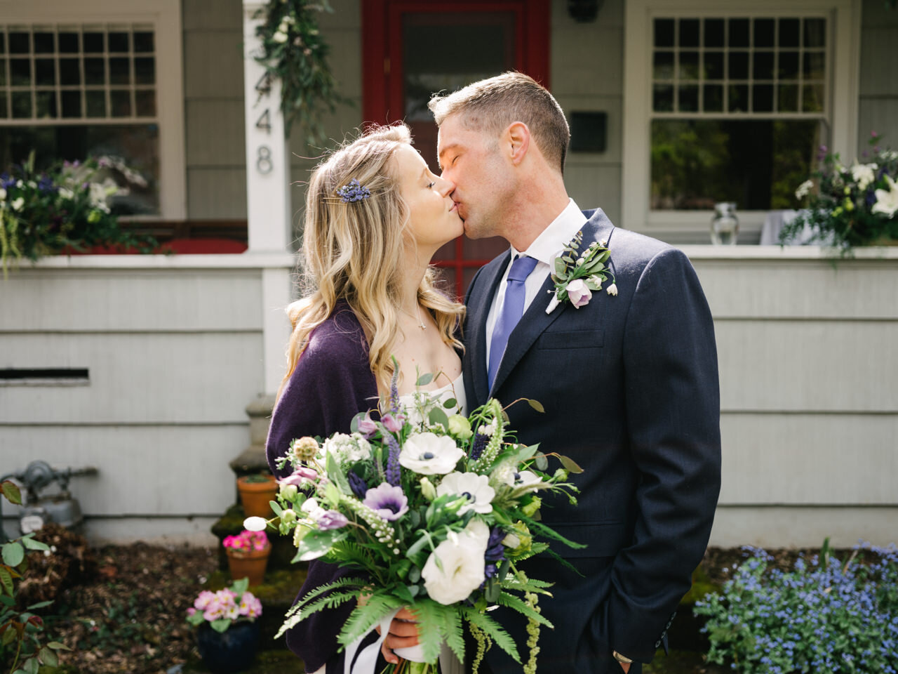 Blonde bride and groom kiss in front of home holding purple, white bouquet with ferns backlit by sunlight 