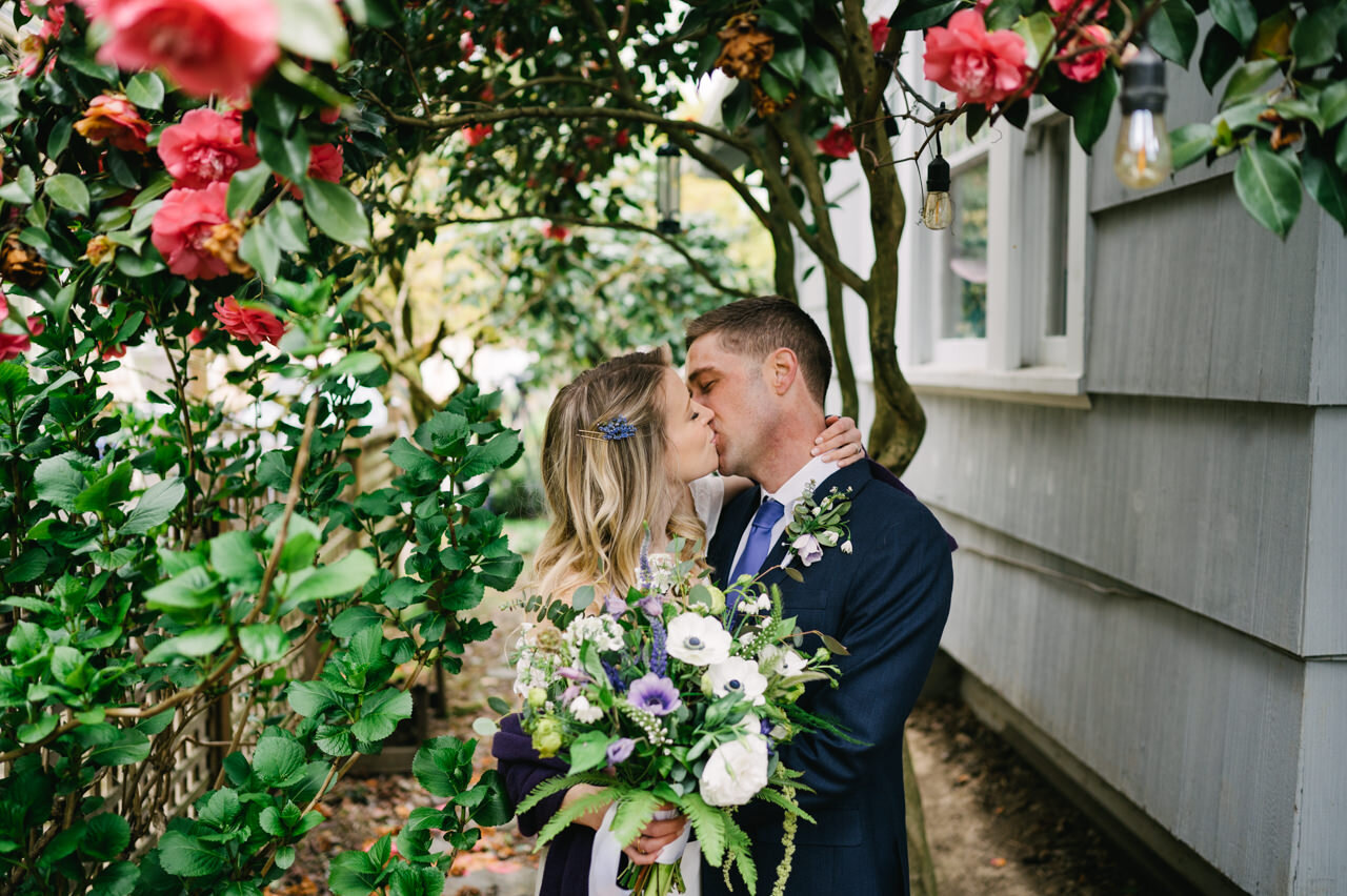  Wedding couple kisses next in house alley under camelia tree with purple and white bouquet 