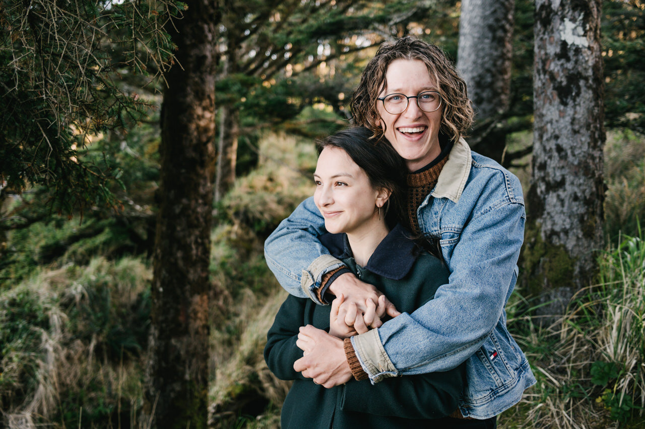  Boy with glasses and long hair smiles while wrapping arms around his fiancé 