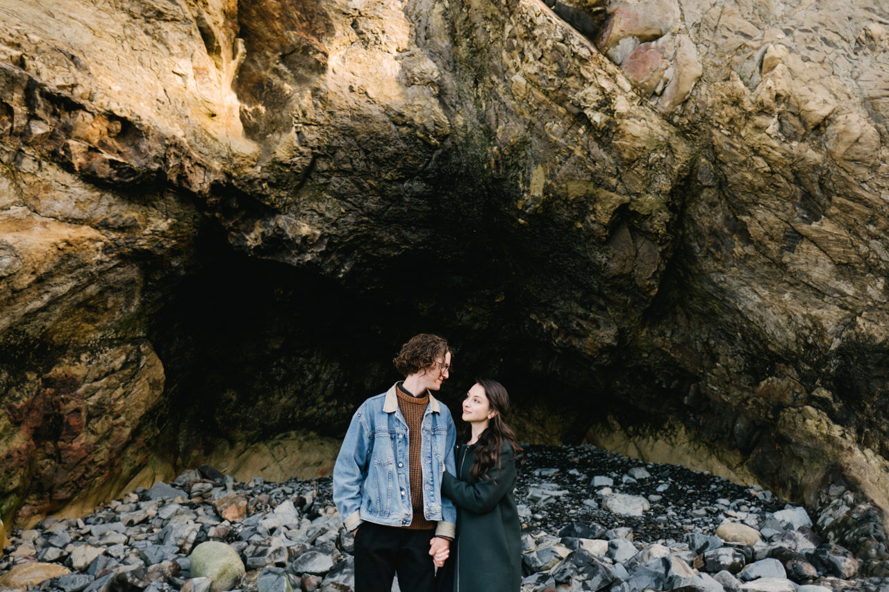 Brown haired woman hugs fiancé arm under rock cave on Oregon coast 