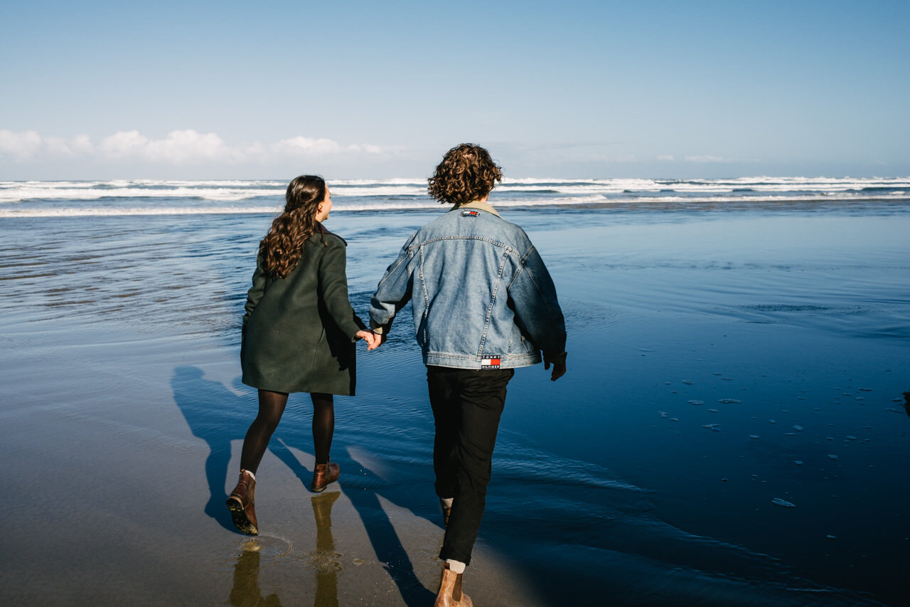  Engaged couple runs around hug point in front of blue ocean 