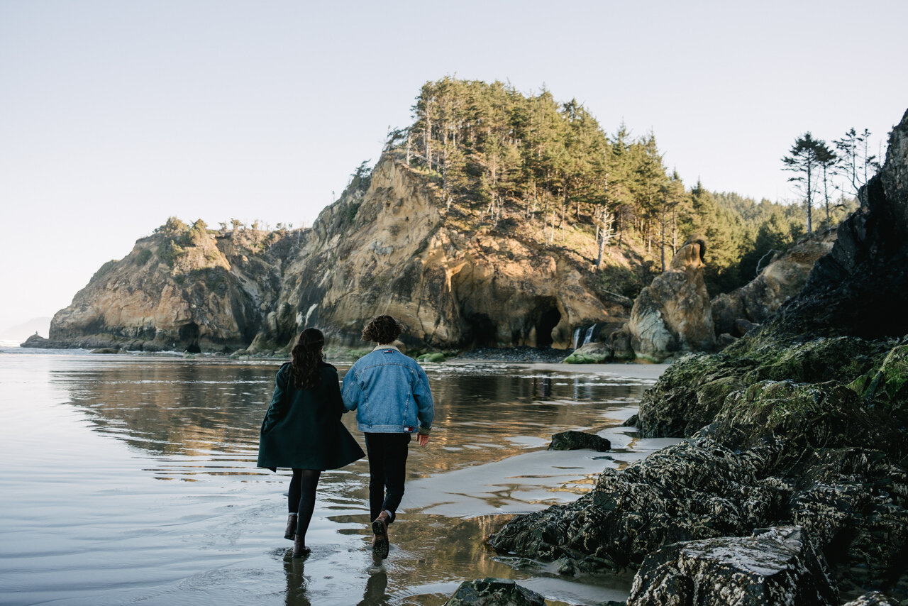  Engaged couple walks toward hug point waterfall in morning sunlight 