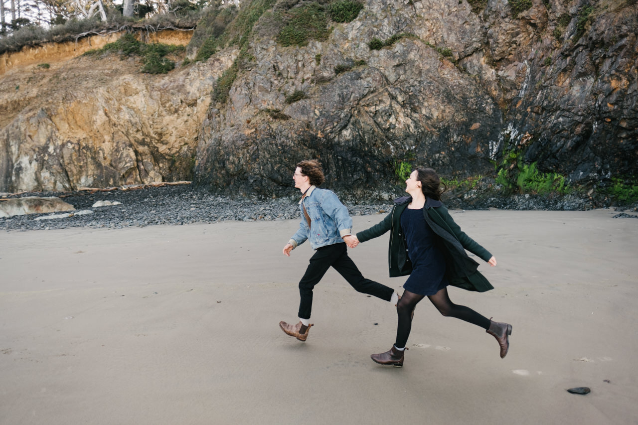  Couple holding hands and running in sand along rocky Oregon coastline 