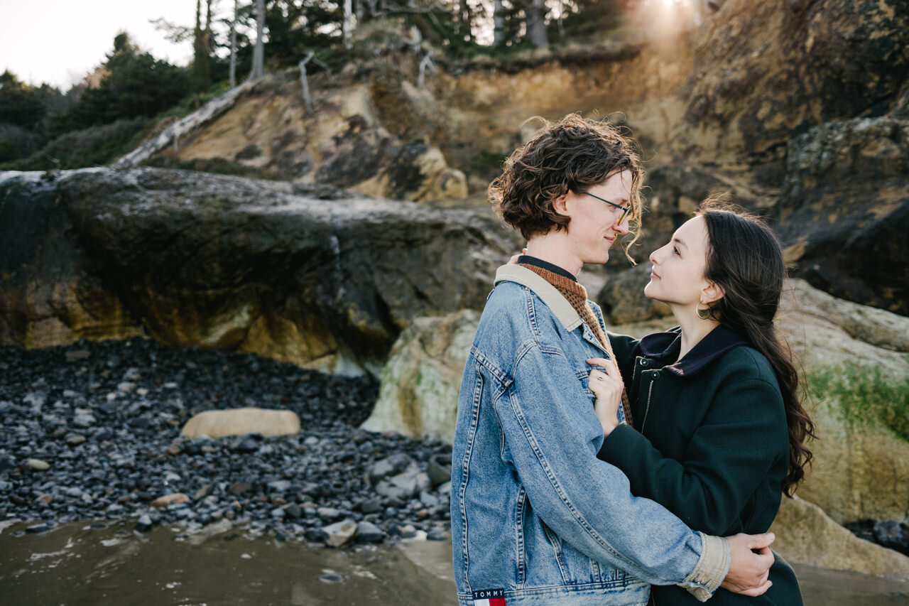  Boy in jean jacket embraces girl in green jacket with morning sunlight peaking over the Oregon coastal forest 