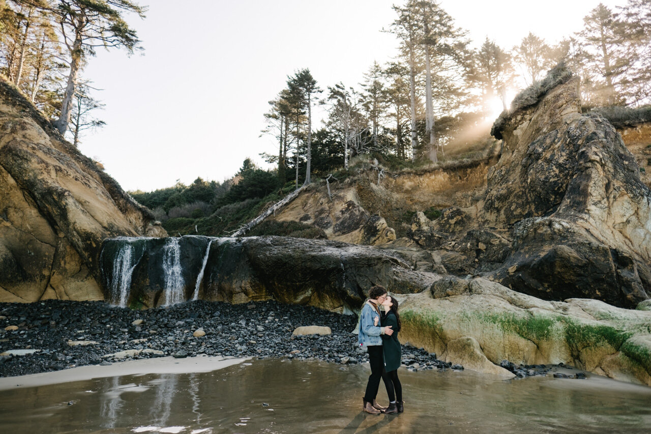  Morning sun rays stream through Oregon coastal forest onto waterfall while engaged couple stands together on fresh wet sand at Hug point 