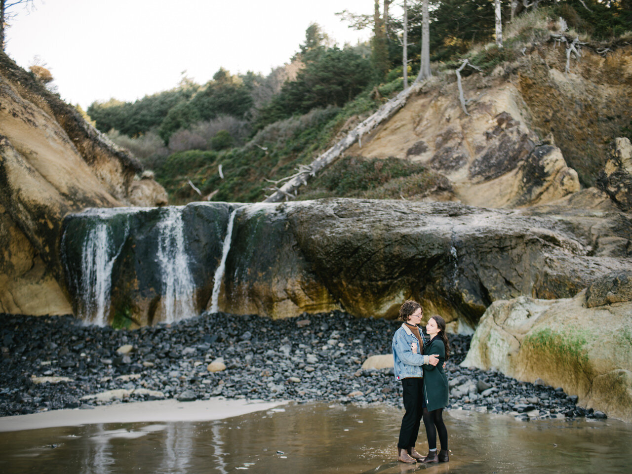  Couple talks together in front of Oregon Hug Point waterfall during engagement session 