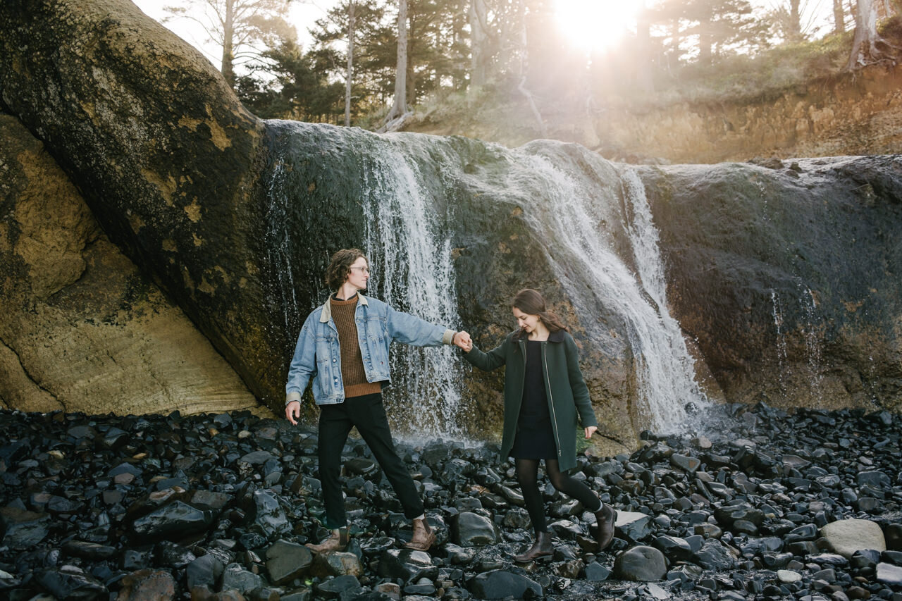  Couple holding hands walks across wet stones in front of waterfall and streaming sunlight 