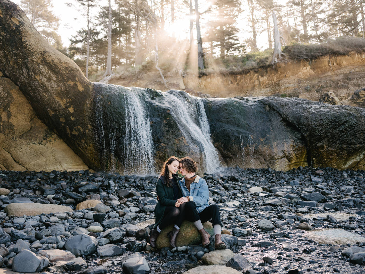  Engagement session in front of hug point waterfall with streaming sun rays behind brown Oregon cliffside 