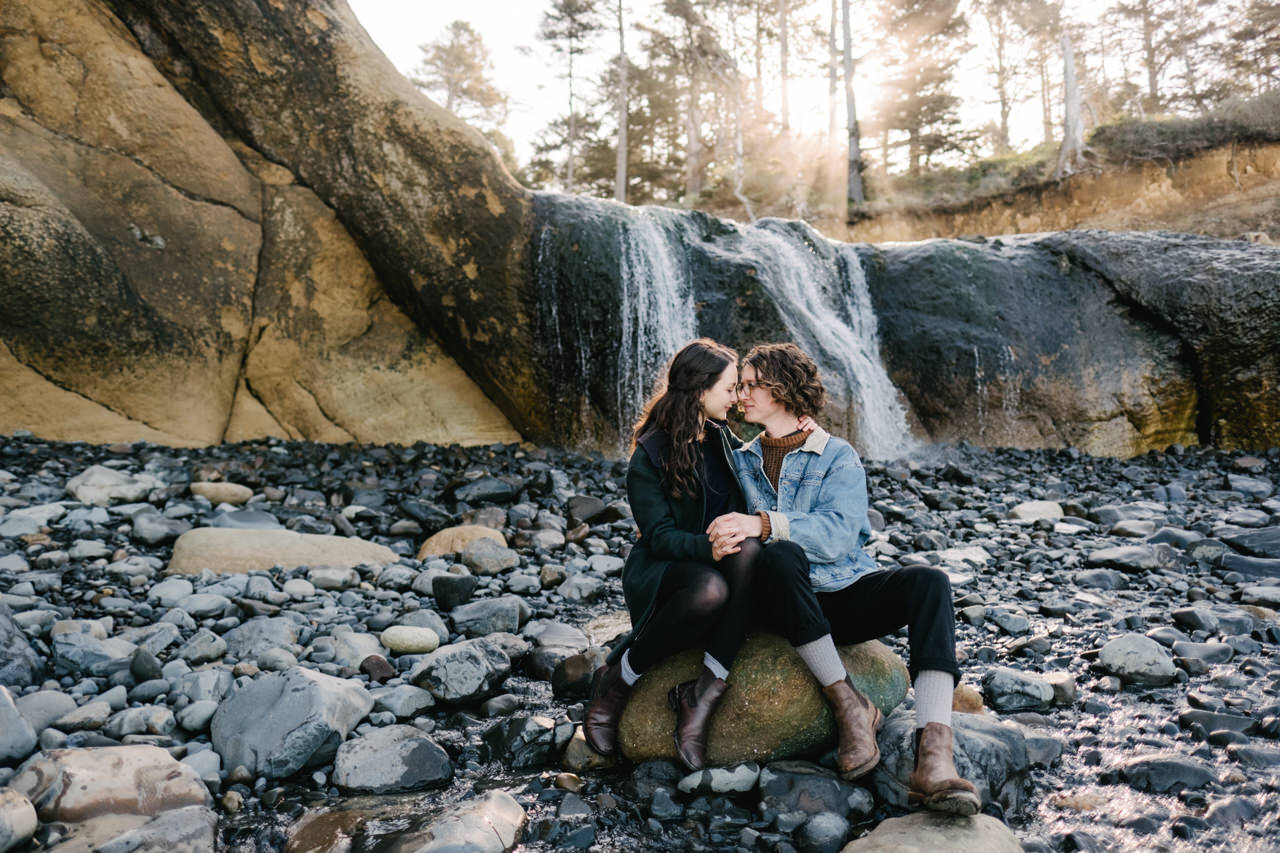  Couple in brown boots and jean jacket sitting on rock in front of Oregon coast waterfall in morning sunlight 