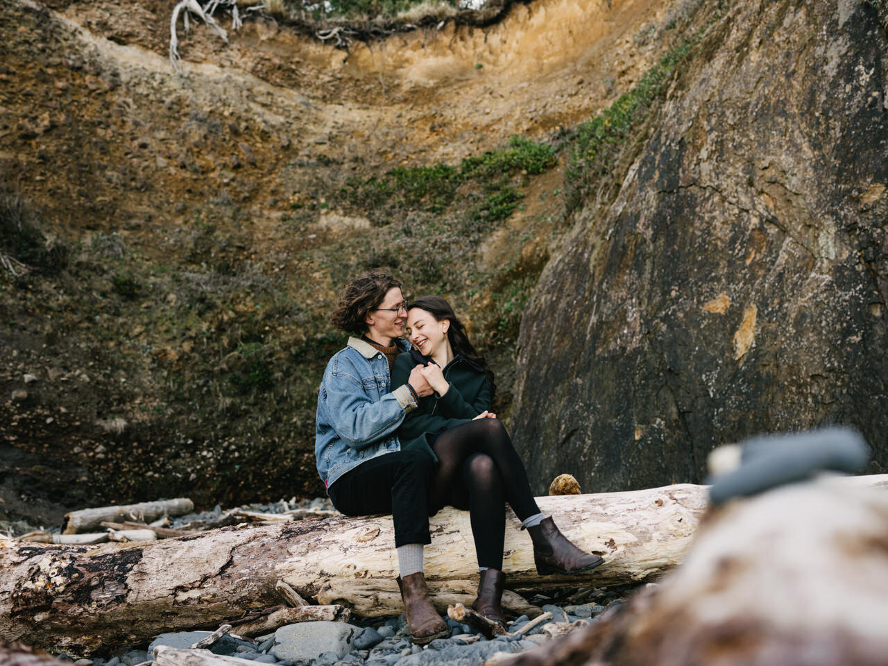  Funny moment while couple sits in front of brown Oregon coast cliffside while sitting on driftwood log 