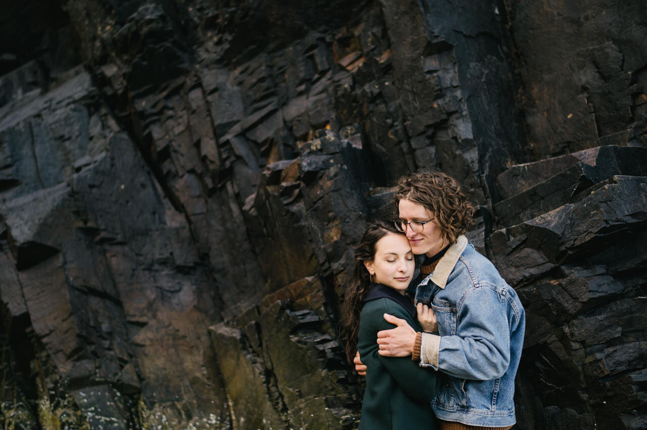  Couple embrace in cold wind in front of coastal rocks 