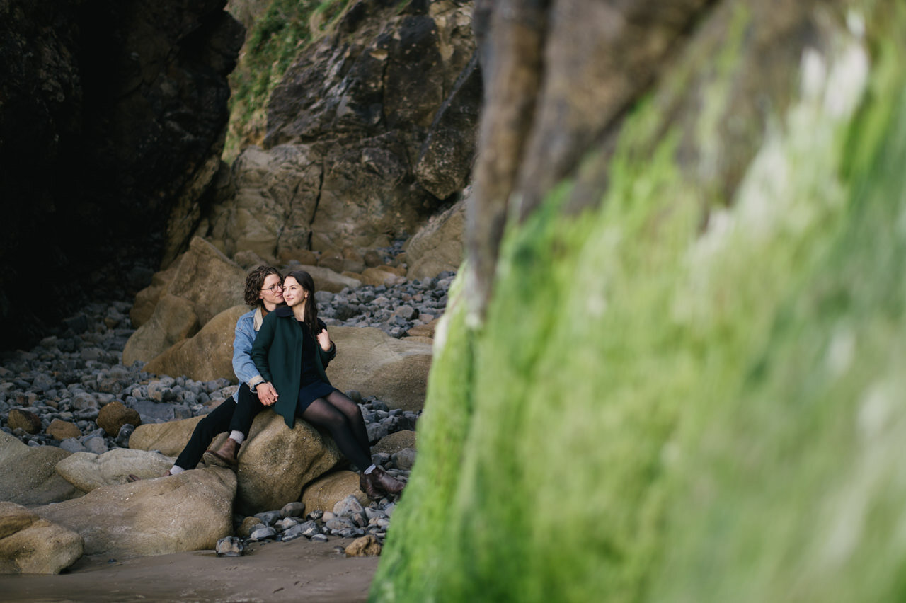 Couple sits together on large rock with wet green ocean algae in the foreground 