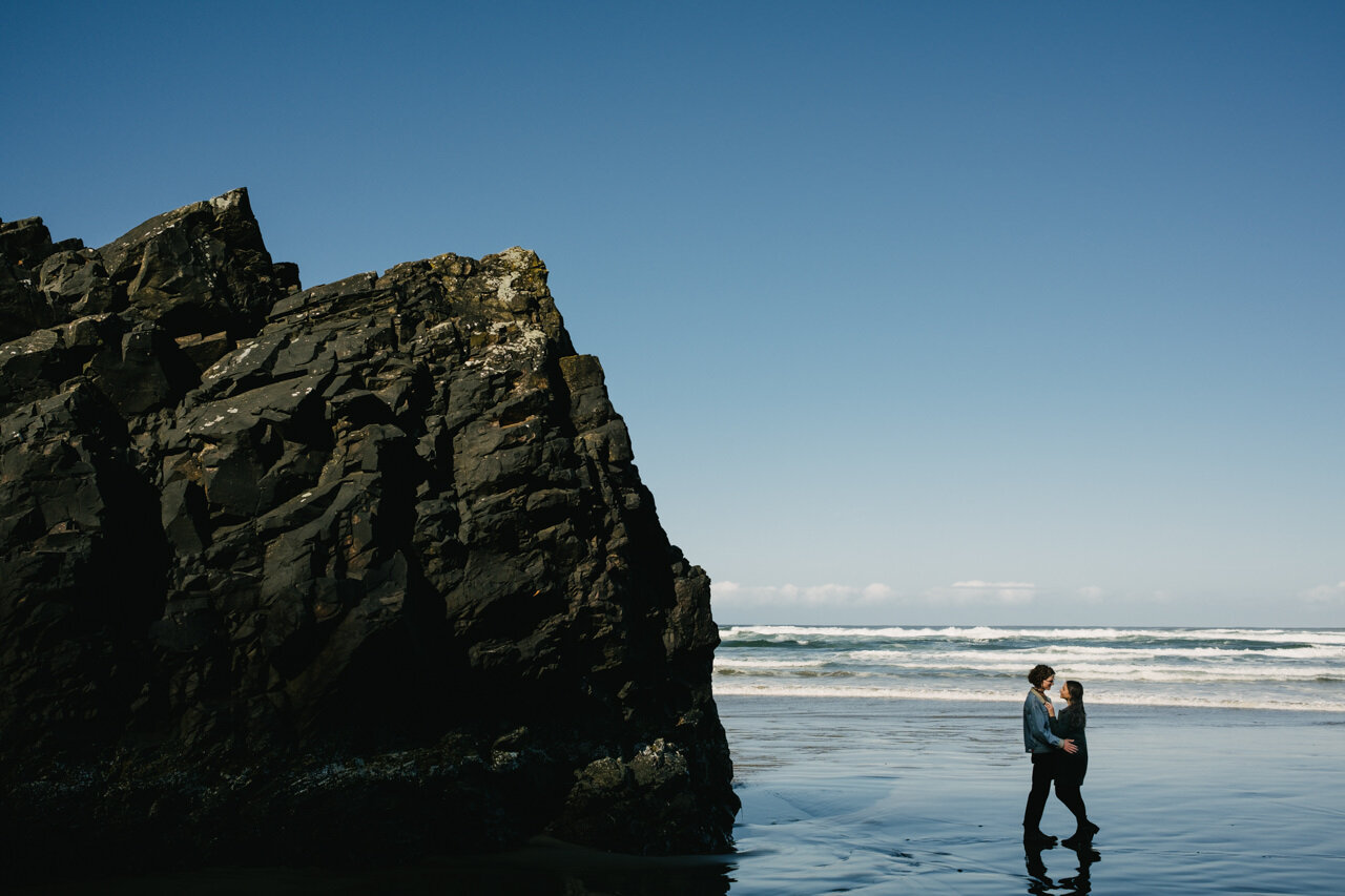  Engagement photo in front of blue ocean and sky with jagged sunlit Oregon coast rock 