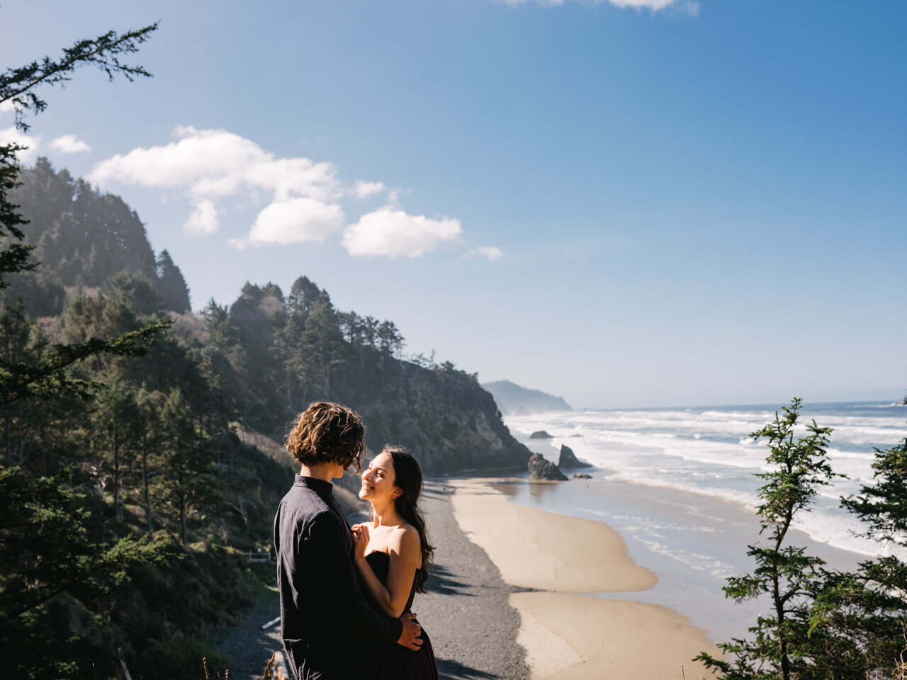  Engaged couple stands on cliffside overlooking sunny Oregon coastline 