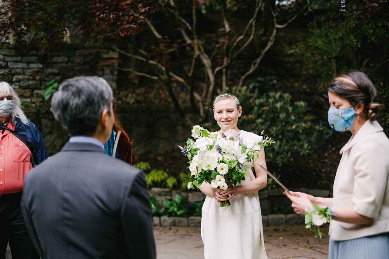  Bride smiles at groom while she holds large white bouquet next to officiant with light blue floral mask 