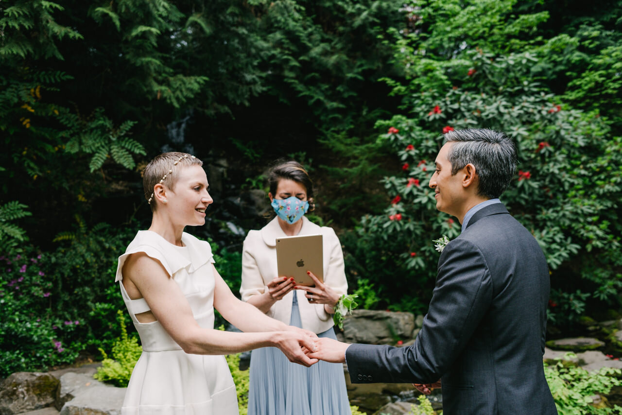  Bride slides ring on grooms finger in front of red Rhododendrons and waterfall 