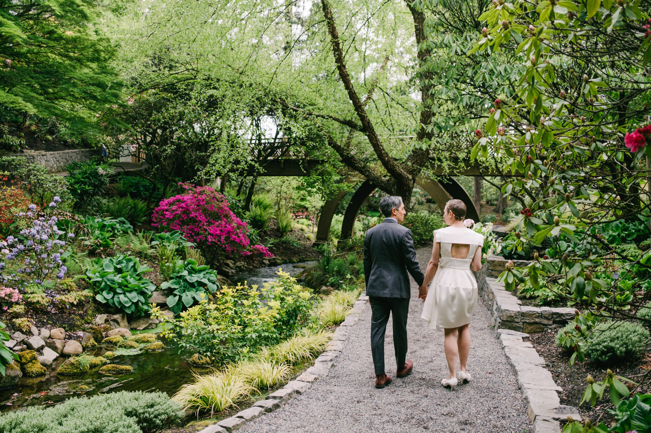  Wedding couple walks down path surrounded by lush gardens of pink azaleas, willow trees and purple flowers 