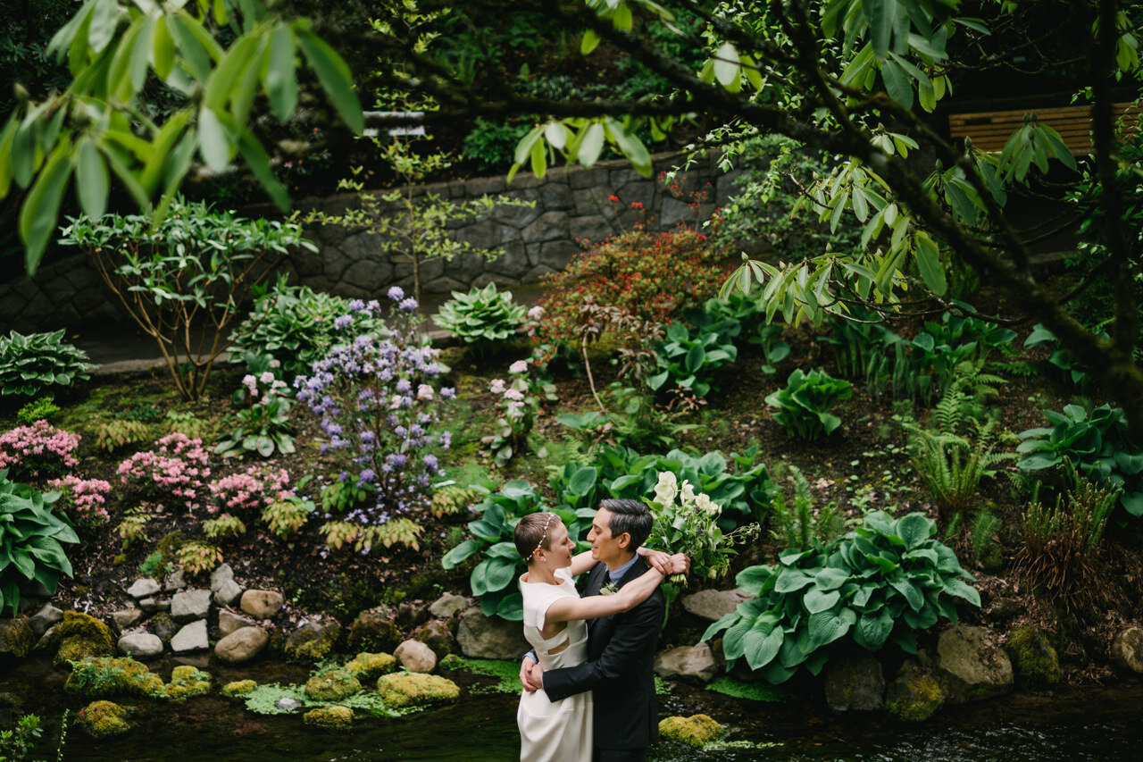  Bride and groom embrace in front of lush stream and pink and purple flowers 