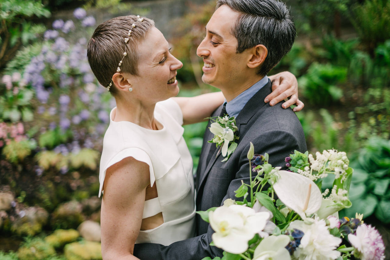  Short haired bride and groom with grey hair embrace around flowers 