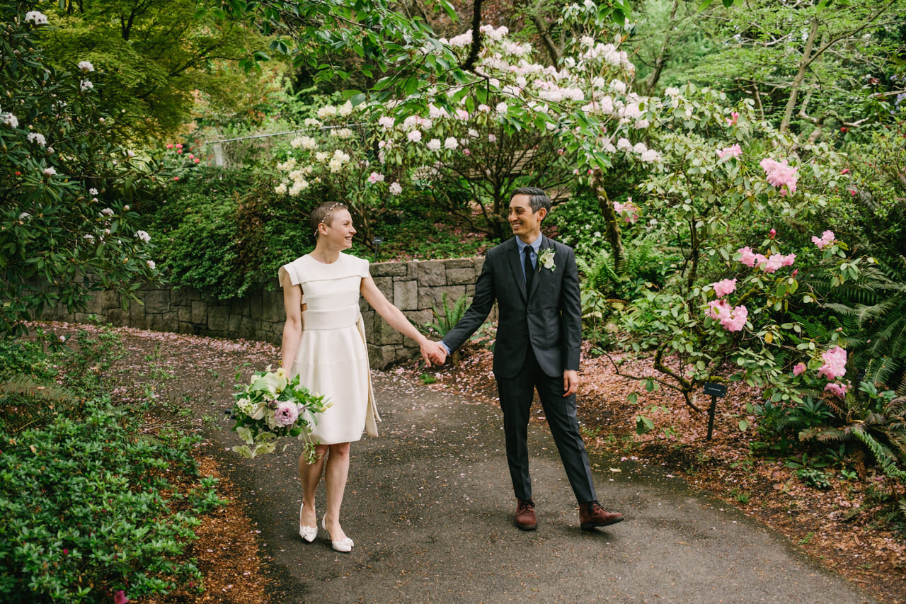  Groom in grey suit and brown shoes hold hands with bride with short white dress walking in gardens 