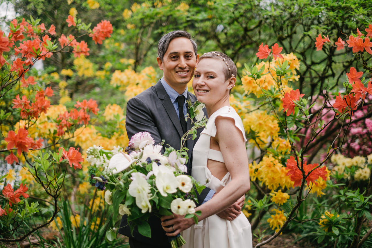  Orange and yellow rhododendron flowers surround bride in white styled dress and groom 