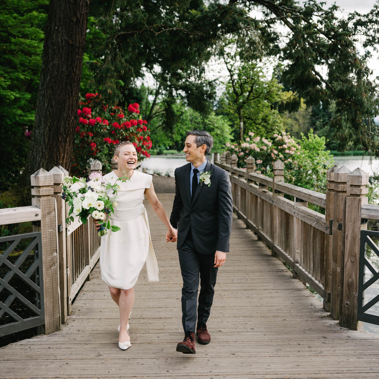  Holding hands across crystal springs bridge, bride laughs while holding large white bouquet 