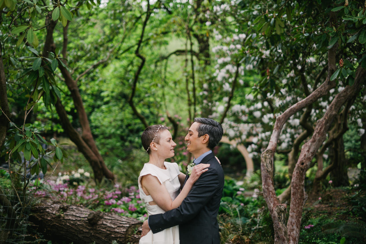  In grove of rhododendrons, groom and bride talk by logs and old bushes 