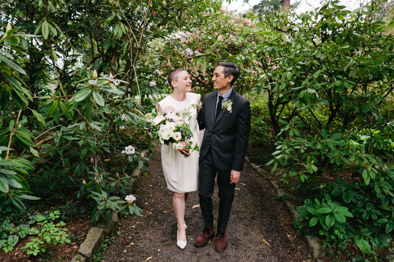  Candid moment of bride and groom walking through Rhododendron bushes in short white dress 