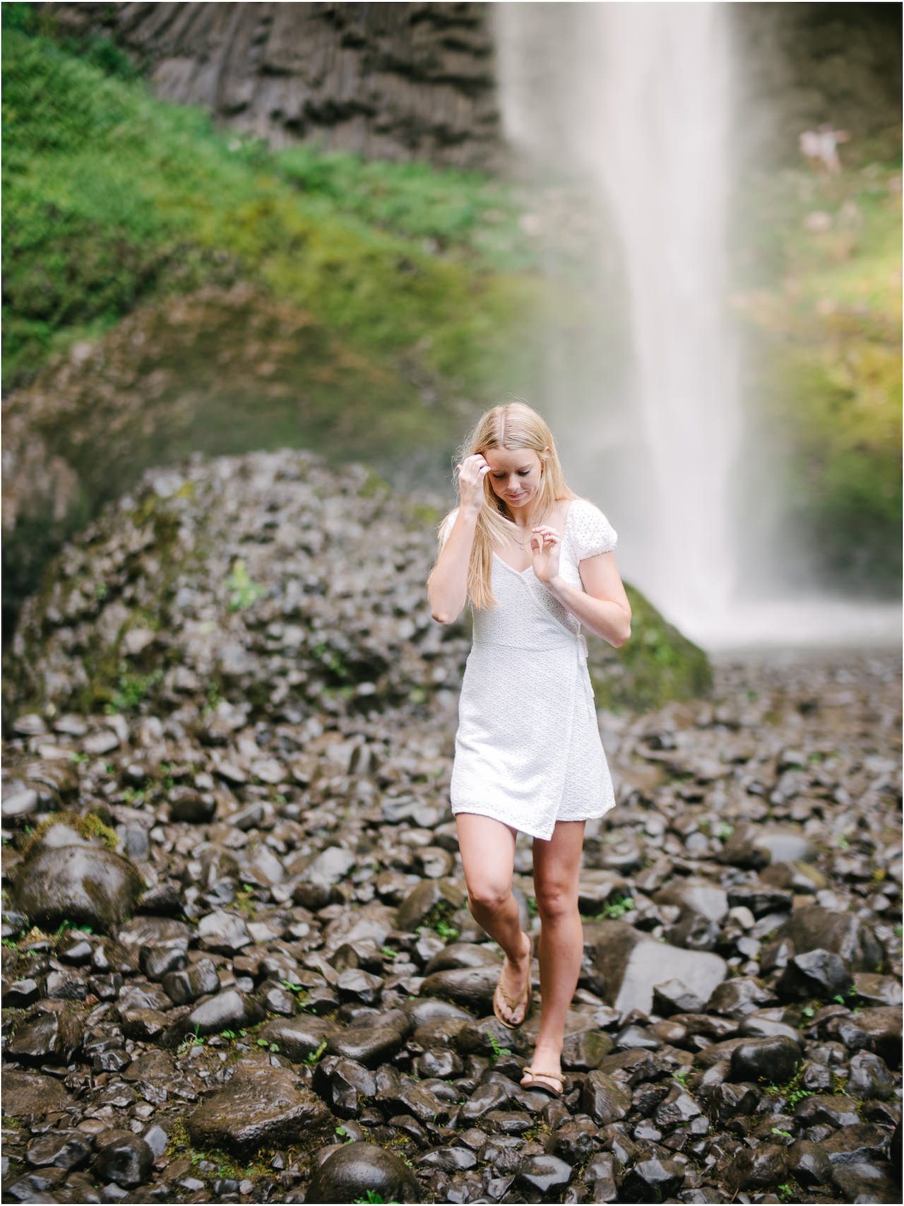  Tall blonde high school student in white dress walks on wet stones away from large waterfall 
