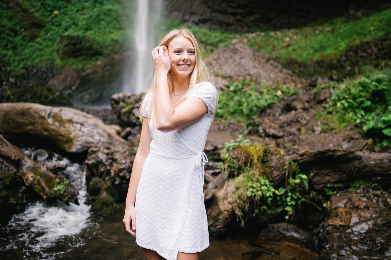  Smiling happy blonde high school senior brushes hair away while standing in creek 