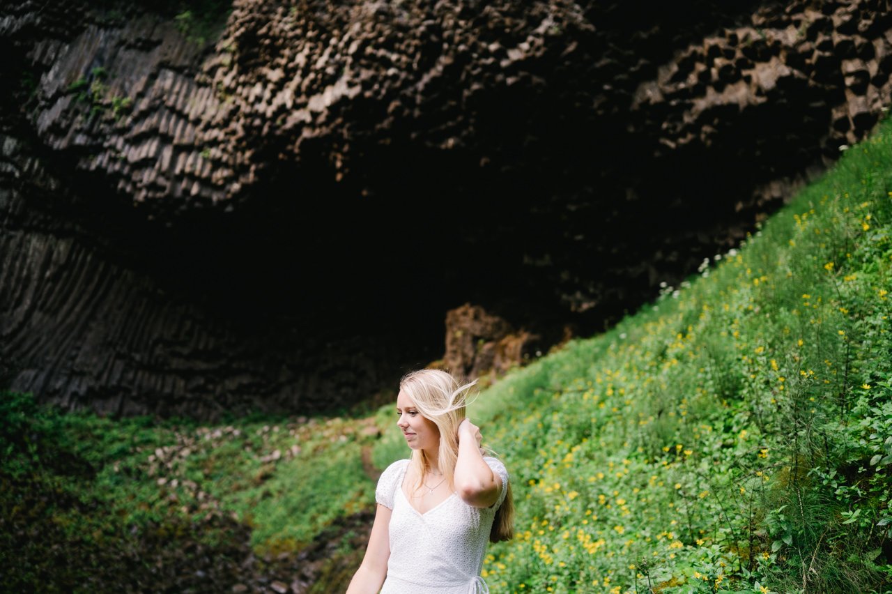  Wind blows hair of blonde senior high school student with basalt rock and green foliage in background 