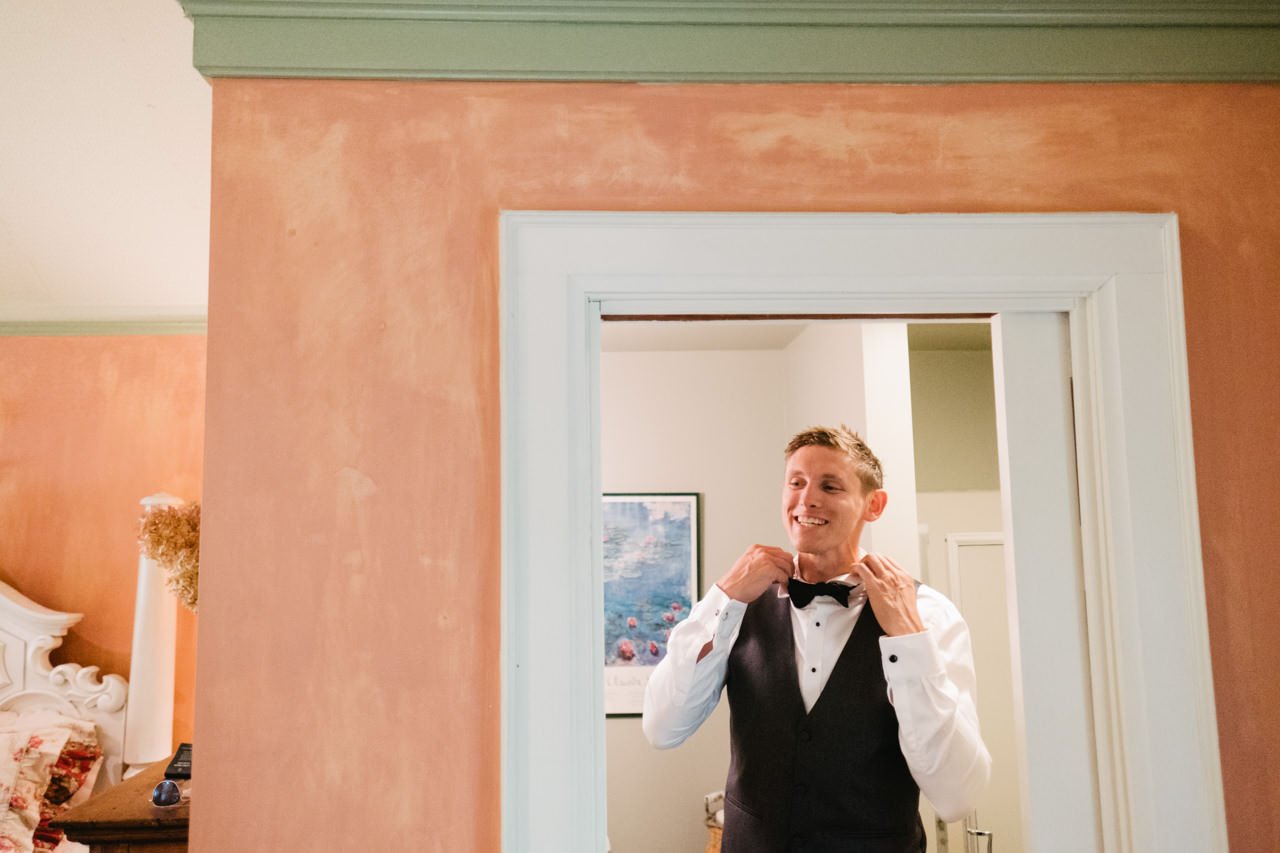  Groom framed in orange walled doorway adjusts bow tie while smiling 