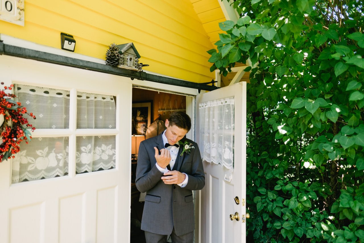  Green foliage encroaches on yellow house where groom adjusts cufflinks 