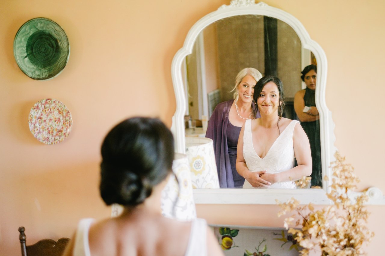  Bride looks at her bridesmaids through mirror while mother in purple dress looks on in orange room 
