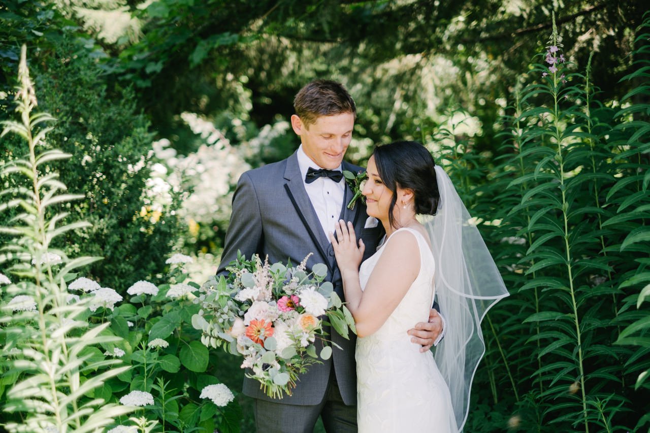  Groom holds orange pink and white bouquet while standing in tall gardens 