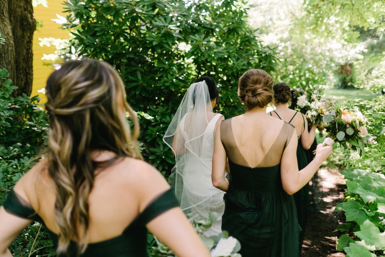  Bride and bridesmaids in green dresses walk away through rhododendrons 