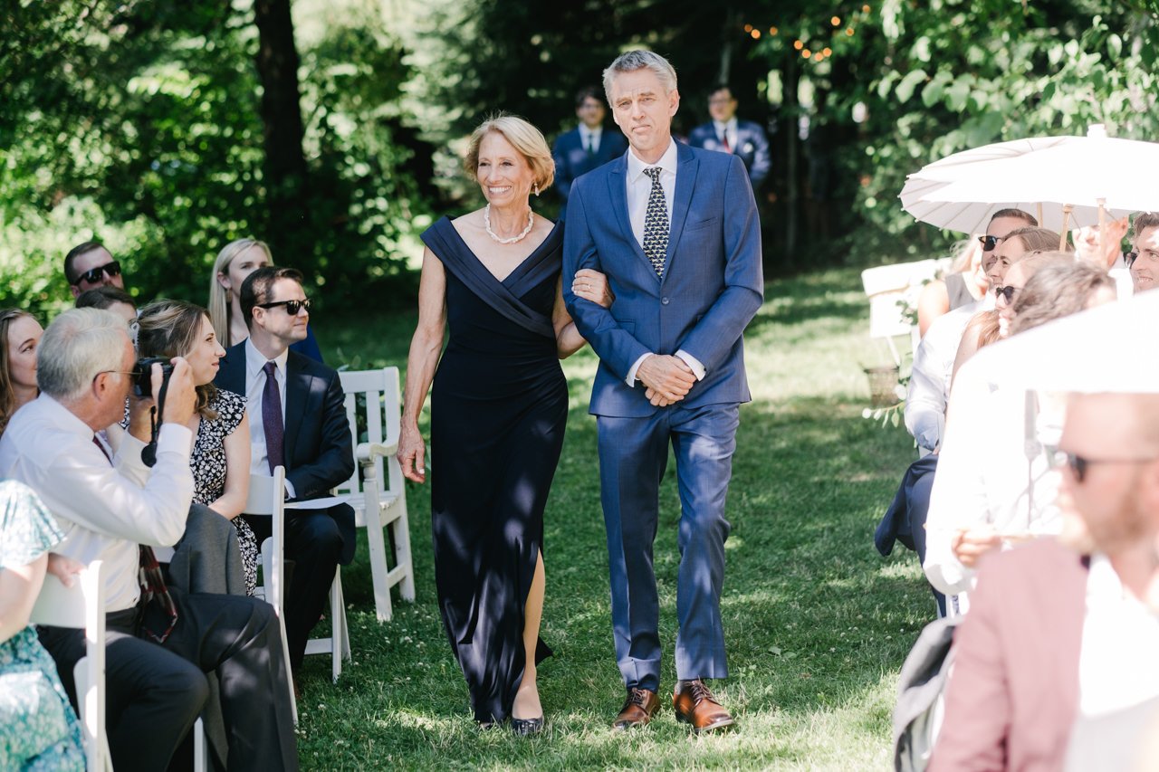  Mother and father of groom walk down aisle in sunlight 