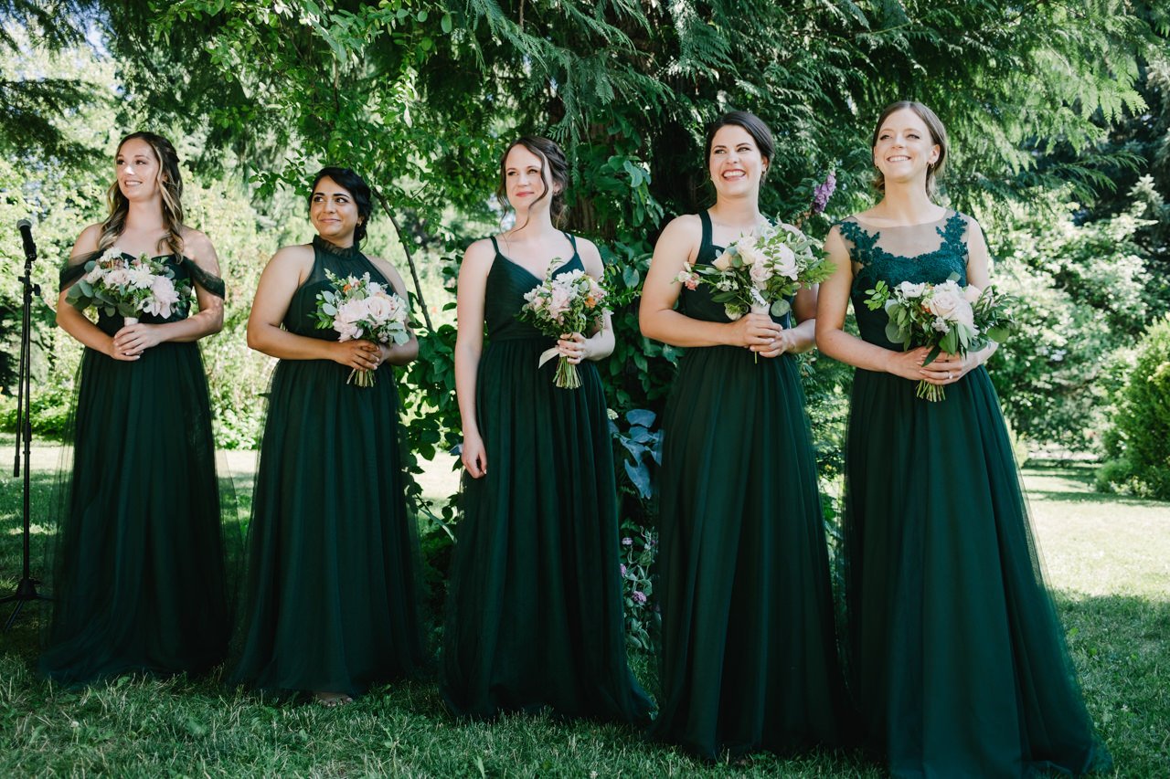  Bridesmaids in green dresses in front of green trees smile as bride approaches 