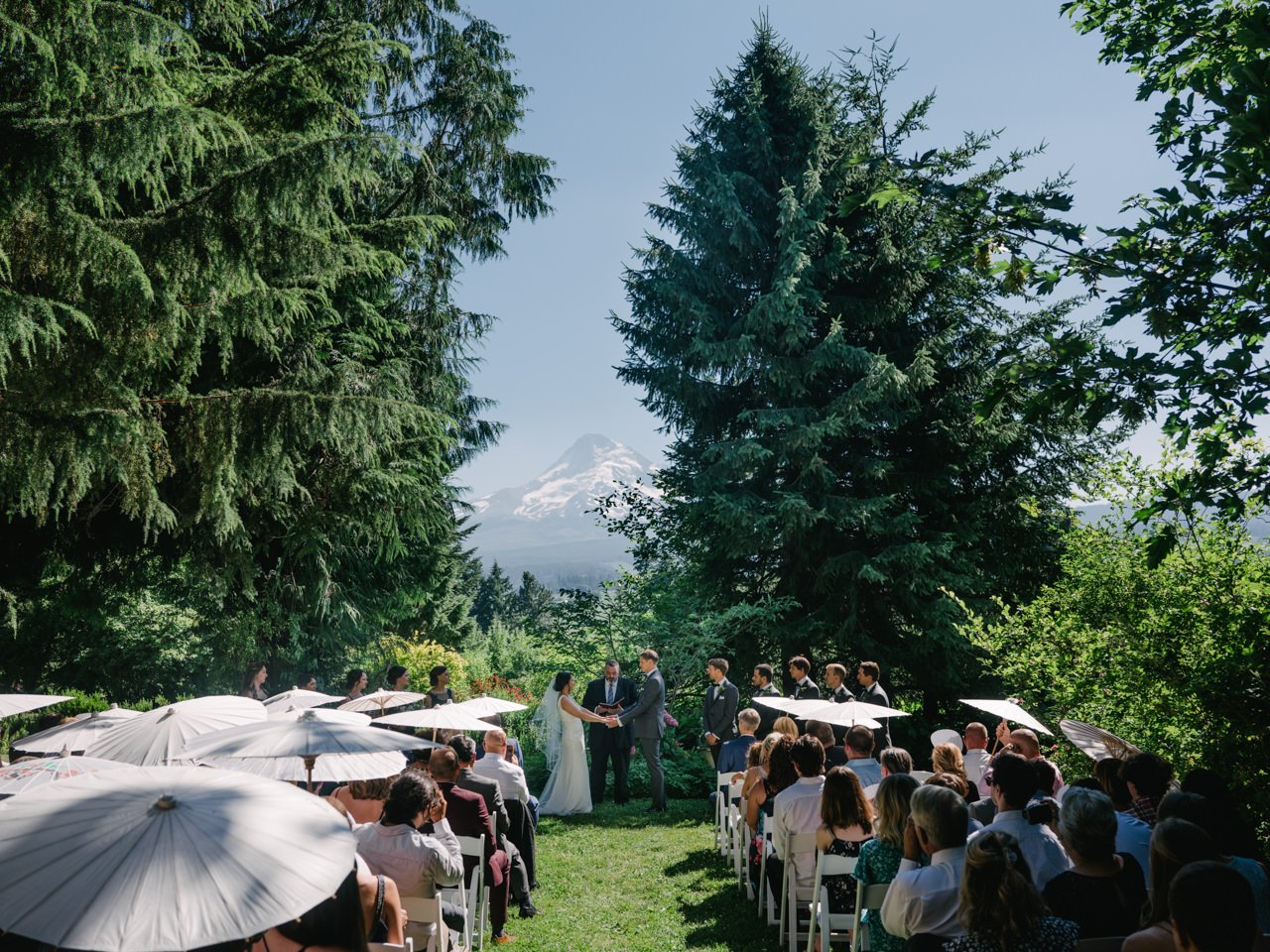  Wedding ceremony at mt hood organic farms in sunshine with guests holding umbrellas in front of mt hood 