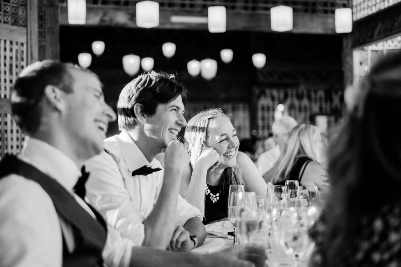  Three wedding guests laughing during toasts with lantern lights of apple house behind them 