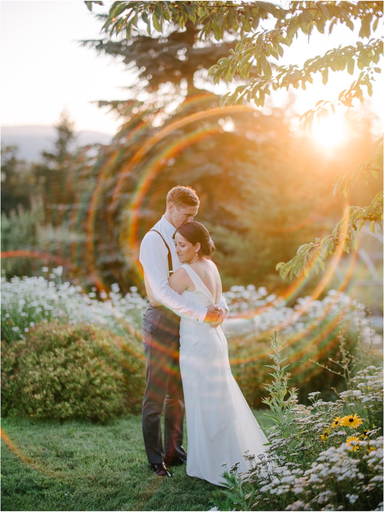  Bride and groom embrace in sun flare in wildflowers in orange evening sunlight 
