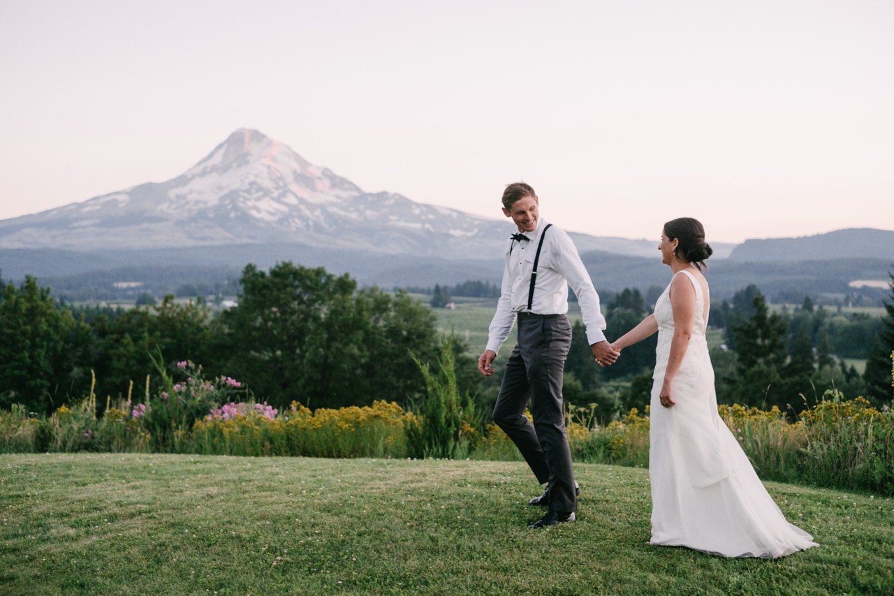  Bride and groom holding hands walk in grass with mt hood and hood river valley behind them 