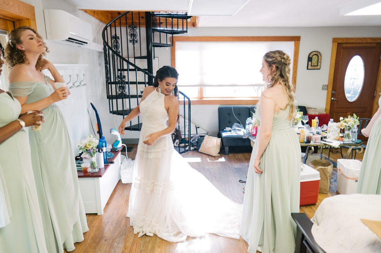  Bride looks at dress in room sunlight surrounded by bridesmaids in light green dresses 