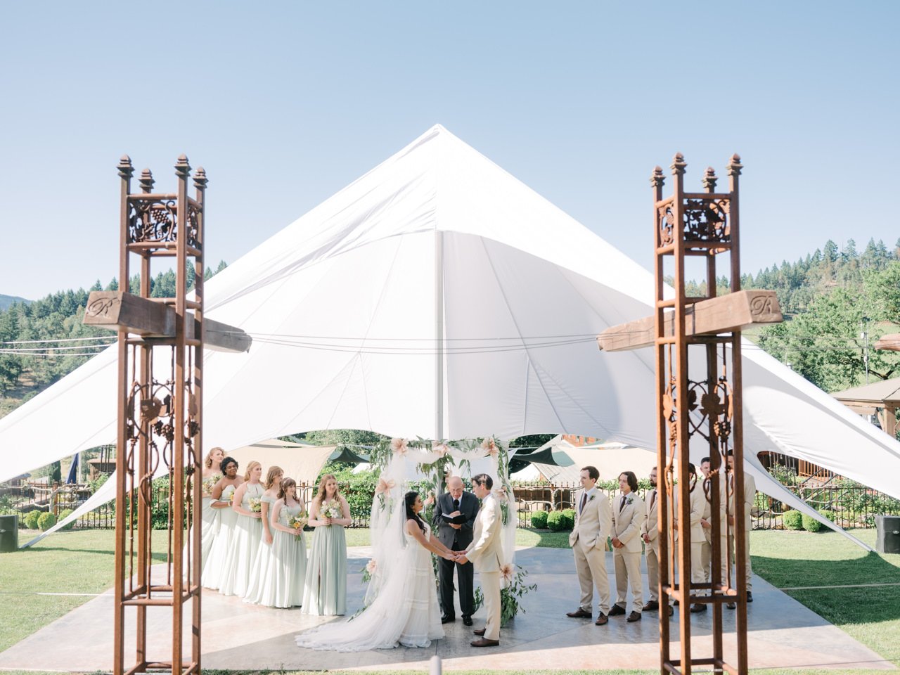  Bride and groom and wedding party face each other under sunlight tent and pink floral backdrop at reustle prayer rock vineyard 